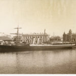 Image: A nineteenth-century steamship with twin funnels and two masts moored at a wharf on the waterfront of a port town. A handful of large nineteenth-century warehouses and other buildings are visible along the waterfront