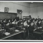 Image: A group of Caucasian men and women dressed in early twentieth century attire practice typing in a classroom. A moustachioed male Caucasian instructor looks on in the background