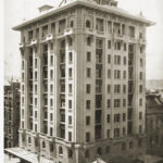 Image: an eleven storey high rise with a hanging verandah from the first floor shading the footpath. Balustrades and small columns adorn the top floor of the building.