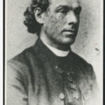 Image: A photographic head-and-shoulders portrait of a young Caucasian man in a Victorian-era Catholic clergyman’s outfit. The man has long, dark wavy hair pulled back behind his head