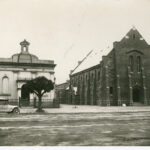 Image: Two Church buildings, old and new are side by side. The smaller old building is topped by a belfry. In front of it is a small tree. A tourer car is parked by the kerb. The larger building is made of dark stone, has a plain facade and butresses.