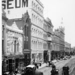 Image: a busy city street lined with three to five storey buildings. There is a single motor car amongst the horse drawn vehicles travelling down the dirt road which also hosts a number of street vendors and their carts.