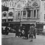 Image: a group of women in 1920s era coats cross an intersection. On the other side of the road are ornate three and four storey buildings with shops including a tailor and Balfours Bakery.