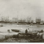 Image: A sketch of the Port Adelaide waterfront during the early nineteenth century. A group of people are engaged in different activities in the foreground, including a man mending nets while a woman looks on. Several ships are moored at a wharf