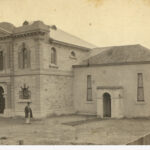 Image: a man stands outside two joined buildings. The newer construction on the left is two storeys and much more ornate than the earlier single storey construction on the right.