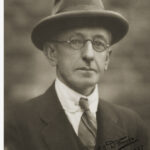 Image: black and white photo of man in glasses, hat and suit