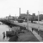 Image: A tram and four pedestrians cross a bridge across a river in a port. A complex of nineteenth-century buildings and a wooden-hulled hulk are visible in the background