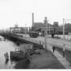 Image: A tram and four pedestrians cross a bridge across a river in a port. A complex of nineteenth-century buildings and a wooden-hulled hulk are visible in the background