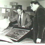 Image: Black and white photograph of two men in suits. They are standing around a just unwrapped painting of a ship