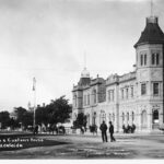 Image: A large, two-storey stone building in Victorian Italianate style. It features a large, octagonal tower with an additional storey in one corner. A horse-drawn dray is present in the street in front of the building