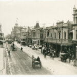 Image: a dirt street, lined with two and three storey stone buildings and busy with horse drawn vehicles, pedestrians and street vendors.