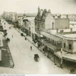 Image: a high angle photograph of a city street lined with shops of various architectural styles, most between two and three storeys high