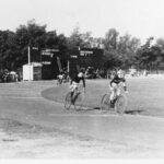 Image: cycle racing at Adelaide Oval. Two men on bicycles vie for the lead on a tarmac track.