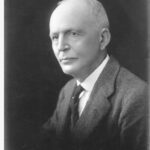 Image: A head and shoulders portrait of a man in three quarter view, silhouetted against a dark background. He is clean-shaven and wears a jacket and waistcoat with a collar and tie.