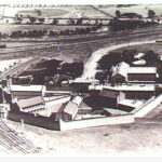 Image: an aerial photograph of a prison arranged in a half octagonal panopticon style. With long rectangular cell blocks inter-spaced with triangular paved yards. A high wall surrounds the buildings.