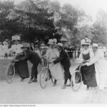 Image: three women on bicycles about to begin a race. Men hold the bicycles steady.