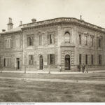 Image: Two men and a boy stand outside a two storey stone building with a corner entrance.