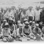 Image: A large group of Aboriginal men, two at the front have tin cans possibly from ration distribution