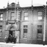 Image: A group of men in early twentieth century attire stand in front of a large, nineteenth century two-storey bluestone building. Signs in the first floor windows read ‘Seamen’s Mission & Sailors’ Rest’