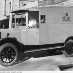 Image: An old motorised van parked next to a building with a man sitting in the front drivers seat