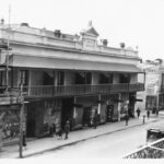 Image: Shops and pedestrians seen from an elevated position on the opposite side of the road. The row of two storey shops in the centre has parapet and a cantilever verandah. The building on the left is covered in scaffolding.