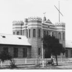 Image: A large, two-storey brick building built in mock-Tudor style, with two turrets flanking a large entrance door. A white picket fence surrounds the building, and a motorcycle with side-car is parked outside the fence under a tree