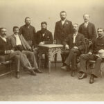 Image: A group of eight young Caucasian men in late Victorian attire and varying styles of facial hair pose for a photograph in a studio. Some of the men are seated on cane chairs around a small ornate cane table. Others stand behind