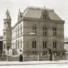 Image: Two women in early 20th century clothing, one with a sun umbrella, stroll past a large stone building with two main floors as well as basement and attic space.