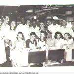 Image: Nineteen men and women in a ship's kitchen. Seven women and one man are seated at a table, the rest stand behind them. They are all wearing white or aprons