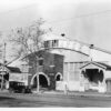 Image: a single storey wooden building with a pitched gable roof and protruding arched brick entrance way. There are 1920s era cars parked outside.