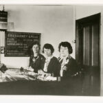 Image: Three women are seated at a switchboard. Behind them is a sign that says "Emergency Calls" and lists numbers for police, fire, ambulance, hospital and waterworks