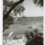 Image: Black and white photograph of people on a beach, with boats in the water in the distance.