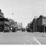 Image: a city street with a mixture of horse drawn and motor vehicles. The street is lined with shops.