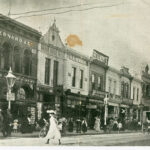 Image: a woman in a white dress crosses a road to join a large number of other pedestrians walking on the footpath. The road is lined with two storey terraced shops. A policeman stands under a street light to the left of the image.