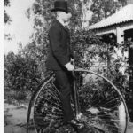 Image: man in a top hat and suit seated on a penny-farthing bicycle