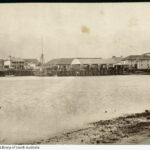 Image: A mid-nineteenth century landscape photograph of a port waterfront. Several buildings are visible next to the water, and a small number of sailing ships are moored alongside a large wharf