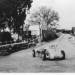 Image: A 1950s vintage racing car drives around a corner and through the streets of a small rural town. A handful of small historic stone buildings are visible on either side of the road