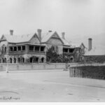 Image: A large two storey house with balconies set behind a stone wall and covered with ivy