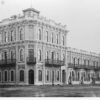 Image: a large, stone, three storey corner hotel with adjoining two storey terrace houses. The windows on both the hotel and houses are arched and the roofs flat. The houses have bay windows on the first floor and balconies on the second.