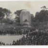 Image: A large crowd of people in 1930s dress or military uniforms gather around a huge stone monument featuring three bronze statues of a girl, a student and a farmer in front of a large marble angel.