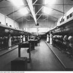 Image: A telephone exchange. There are two rows of switchboard operators on either side of the image, separated by a floor and desks.