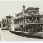 Image: Black and white photograph of two paddlesteamers on a river. Each paddlesteamer has three levels and people are visible standing and sitting on each one. The foremost paddlesteamer is flying the Union Jack flag.