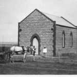 Image: A Caucasian man in a hat and light-coloured top stands outside the front door of a small Bluestone chapel in a remote setting. A horse and buggy stand nearby