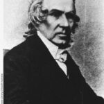 Image: A head-and-shoulders portrait of a middle-aged Caucasian man in mid-nineteenth century clerical attire. He is clean-shaven and has slightly long, shaggy greying hair