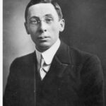 Image: A photographic head-and-shoulders portrait of a young man wearing a suit and wire-rimmed spectacles