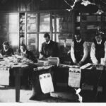 Image: group of men in suits and waistcoats standing around a long table