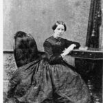 Image: woman in long dress sitting on a chair and leaning on a table.