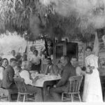 Image: Men and Women around a table underneath a brush and eucalyptus shelter