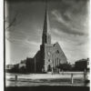 Image: a simple stone church at the intersection of two wide roads. The tower, which is to one side of the building is square at ground level and topped with crenellations. Above this the tower becomes octagonal and is topped with a very steep roof