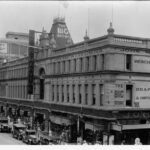 Image: A three storey department store on a busy city street. A large number of 1930s era cars are parked outside and the sidewalk is busy with people, particularly women. Signs on the building identify it as "The Big Store"